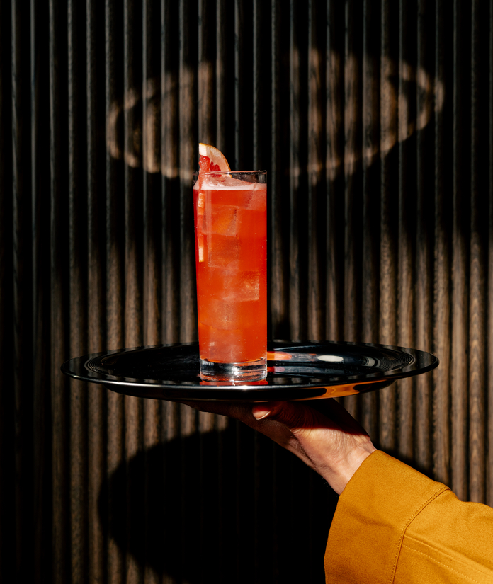 Hand holding black tray with a vibrant pink drink garnished with a citrus slice against a dark textured background.
