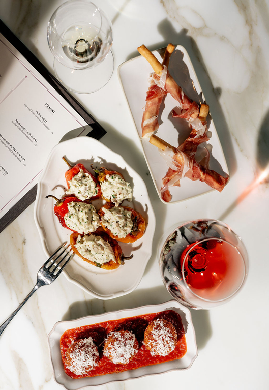 A marble table setting with tapas, including stuffed peppers, meatballs, prosciutto-wrapped bread sticks, and a glass of red wine, next to a menu.