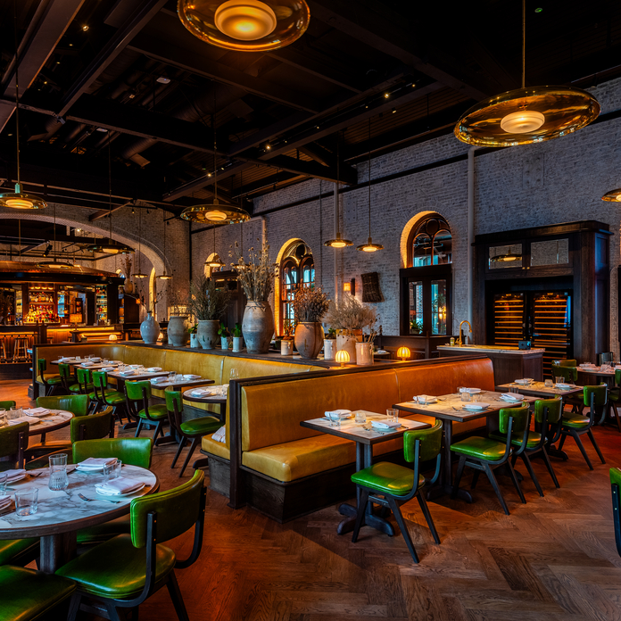 Modern restaurant interior with wooden tables, green chairs, arched windows, and exposed brick walls.