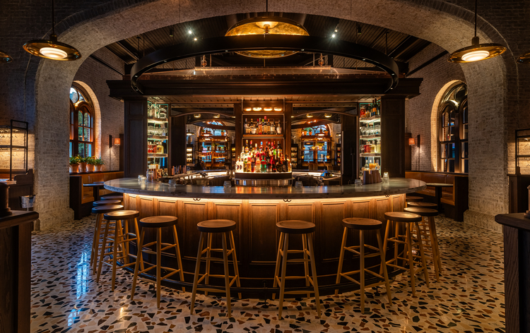 Modern bar with curved counter, wooden stools, stone floor, and metallic accents.